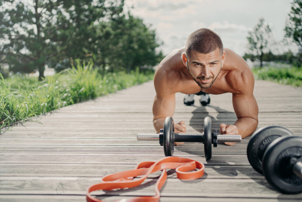 Strong motivated handsome man stands in plank pose, makes abdominal exercises, poses near sport equipment, listens music in wireless earphones, has workout outdoor, looks with serious expression